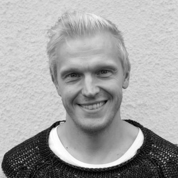 Hymyilevä mies, jolla on lyhyet vaaleat hiukset ja jolla on tumma villapaita valkoisen paidan päällä, seisoo vaalean seinän edessä. Kuva on mustavalkoinen.