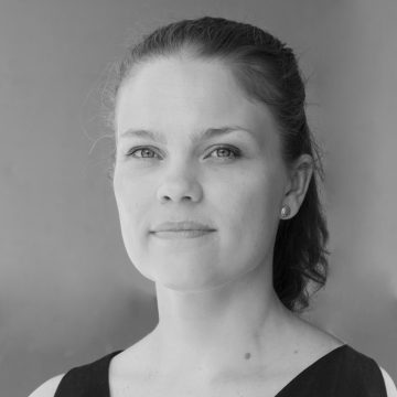 Kameraan katsoo vaaleaihoinen nainen, jolla on vaaleat silmät, hihaton toppi ja pyöreät korvakorut. Hänen hiuksensa on vedetty taakse, ja hänen ilmeensä on neutraali. Tausta on tavallinen ja pehmeästi valaistu.