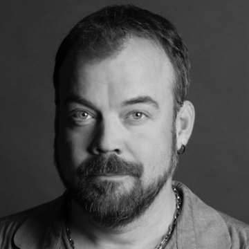 Mustavalkoinen muotokuva parrakas mies, jolla on lyhyet hiukset, päällään ketjukoru, paita ja pieni korvakoru vasemmassa korvassa, katsoo suoraan kameraan neutraali ilme.