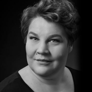 Mustavalkoinen muotokuva henkilöstä, jolla on lyhyet, aaltoilevat hiukset, vaaleat silmät ja neutraali ilme, yllään tumma toppi, tummaa taustaa vasten.