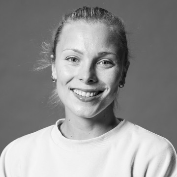 Nuori nainen, jolla on vaaleat taakse vedetyt hiukset, hymyilee kameralle. Hänellä on vaalea collegepaita ja pienet korvakorut. Tausta on yksinkertainen ja harmaa. Kuva on mustavalkoinen.