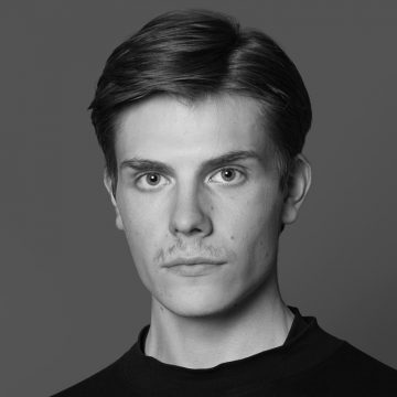 Mustavalkoinen muotokuva nuoresta henkilöstä, jolla on lyhyet tummat hiukset, heikot viikset ja neutraali ilme, yllään tumma kauluspaita, harmaata taustaa vasten.