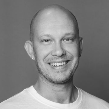 Tässä mustavalkoisessa muotokuvassa hymyilevä kalju mies, jolla on vaalea iho, valkoinen t-paita ja pienet korvakorut, poseeraa harmaata taustaa vasten.