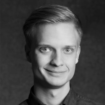 Vaaleahiuksinen ja vaaleasilmäinen nuori mies hymyilee kameralle. Hänellä on napillinen paita, ja hänet on kuvattu mustavalkoisena tummaa, yksinkertaista taustaa vasten.