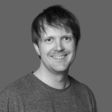 Mies, jolla on keskipitkät, sekaiset hiukset, hymyilee kameralle. Hänellä on yllään yksinkertainen, lyhythihainen, harmaa paita ja hän on poseerannut yksinkertaista harmaata taustaa vasten.