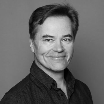 Keski-ikäinen mies, jolla on lyhyet tummat hiukset ja hento hymy, ja jolla on tumma napillinen paita, poseeraa mustavalkoisessa muotokuvassa harmaata taustaa vasten.