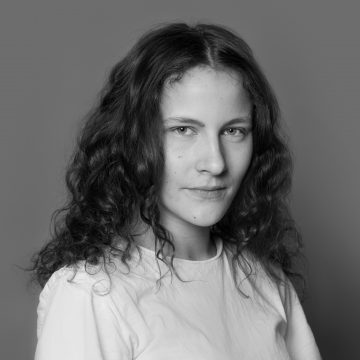 Mustavalkoinen muotokuva nuoresta naisesta, jolla on pitkät, kiharat hiukset, yllään tavallinen vaalea t-paita ja joka katsoo rauhallisesti kameraan neutraalia taustaa vasten.