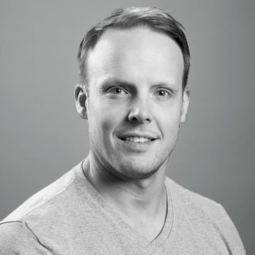 Mies, jolla on lyhyet vaaleat hiukset ja leikattu parta, hymyilee kameralle. Hänellä on yllään vaalea V-kaula-aukkoinen paita ja hän poseeraa vaaleanharmaata taustaa vasten. Kuva on mustavalkoinen.