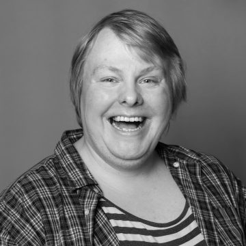 Vaaleahiuksinen henkilö hymyilee leveästi kameralle. Hänellä on yllään ruutupaita raidallisen topin päällä. Kuva on mustavalkoinen ja taustaltaan tavallinen.