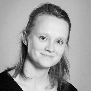 Mustavalkoinen muotokuva naisesta, jolla on vaalea iho, hartioiden pituiset suorat hiukset ja hento hymy. Hänellä on yllään tumma toppi ja hän katsoo suoraan kameraan tavallista taustaa vasten.