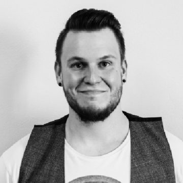 Hymyilevä mies, jolla on lyhyet hiukset ja parta, pukeutuu valkoiseen paitaan ja liiviin. Kuva on mustavalkoinen, ja hän poseeraa tavallisen vaalean seinän edessä.