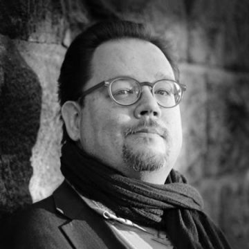 Mustavalkoinen muotokuva miehestä, jolla on silmälasit, lyhyet hiukset ja pukinparta, päällään huivi ja takki, seisoo kiviseinää vasten ja katsoo hieman ylöspäin.