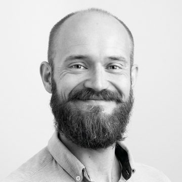 Mustavalkoinen muotokuva hymyilevästä miehestä, jolla on täysi parta ja viikset ja jolla on vaalea nappipaita. Hänellä on taantuva hiusraja ja hän on kameraan päin tavallista taustaa vasten.