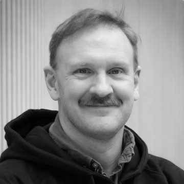 Mies, jolla on lyhyet vaaleat hiukset ja viikset, hymyilee kameralle. Hänellä on tumma huppari kauluspaidan päällä. Tausta on sumea pystysuorilla viivoilla. Kuva on mustavalkoinen.