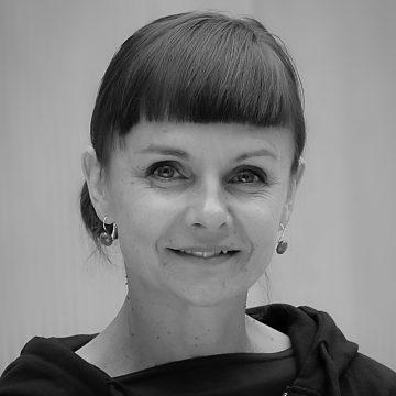 Mustavalkoinen muotokuva naisesta, jolla on lyhyt, suora otsatukka, hiukset taaksepäin vedettyinä, korvakorut ja tumma toppi, hymyilee pehmeästi kameralle tavallista taustaa vasten.