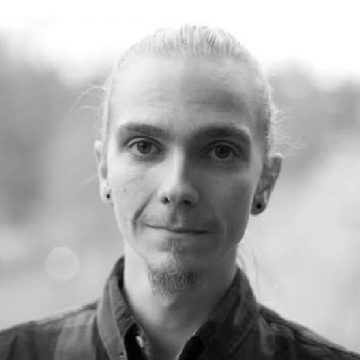 Henkilö, jolla on vaalea iho, taakse sidotut pitkät vaaleat hiukset, lyhyt pukinparta ja pienet korvakorut, katsoo suoraan kameraan neutraalilla ilmeellä. Tausta on pehmeästi sumea.