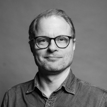 Mustavalkoinen muotokuva keski-ikäisestä miehestä, jolla on silmälasit, vaaleat hiukset ja lyhyt parta. Hänellä on farkkupaita ja hän hymyilee kevyesti kameraan päin tavallista taustaa vasten.