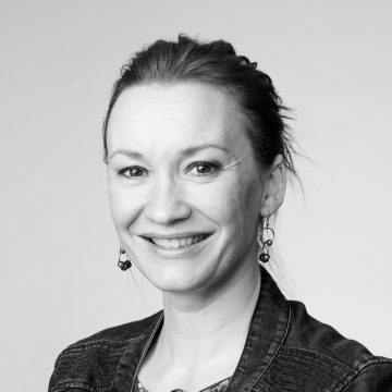 Vaaleaihoinen nainen, jolla on tummat hiukset taaksepäin vedettynä, hymyilee kameralle. Hänellä on yllään roikkuvat korvakorut, farkkutakki ja kuviollinen toppi. Kuva on mustavalkoinen ja taustaltaan tavallinen.