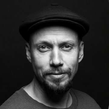 Mustavalkoinen muotokuva miehestä, jolla on litteä lippalakki ja joka katsoo suoraan kameraan hymyillen. Hänellä on lyhyt parta ja viikset, ja tausta on yksinkertainen ja tumma.