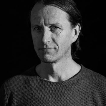 Mustavalkoinen muotokuva keski-ikäisestä miehestä, jolla on suorat, sivulle erotetut hiukset, päällään kuvioitu pusero, katselee hieman vasemmalle neutraali ilme, tumma tausta.