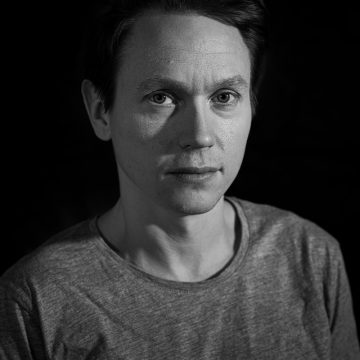 Mustavalkoinen muotokuva henkilöstä, jolla on lyhyet tummat hiukset, yllään tavallinen vaalea paita, katselee suoraan kameraan neutraali ilme tummaa taustaa vasten.