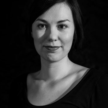 Mustavalkoinen muotokuva naisesta, jolla on lyhyet tummat hiukset, päällään kaula-aukkoinen toppi, katselee kameraan neutraali ilme, tummaa taustaa vasten.