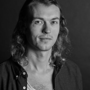 Mustavalkoinen muotokuva nuoresta miehestä, jolla on pitkät, aaltoilevat hiukset, päällään valkoinen t-paita ja avonainen paita, katselee kameraan neutraalilla ilmeellä tummaa taustaa vasten.