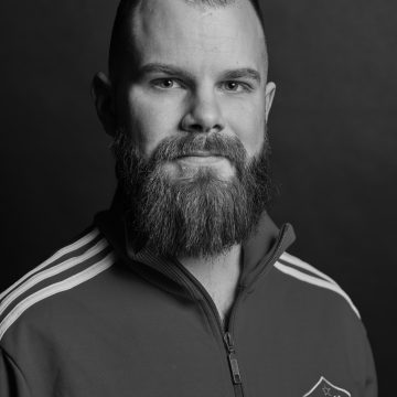 Mustavalkoinen muotokuva parrakkaasta, lyhythiuksisesta miehestä, jolla on yllään Adidaksen vetoketjullinen takki, jonka rinnassa on vaakuna, ja joka katsoo suoraan kameraan tummaa taustaa vasten.