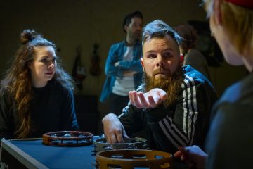 Parrakas mies elehtii kädellään puhuessaan naiselle pöydän ääressä tamburiinien kanssa, kun toinen nainen ja mies katselevat taustalla. Tilanne vaikuttaa epäviralliselta ja vilkkaalta.