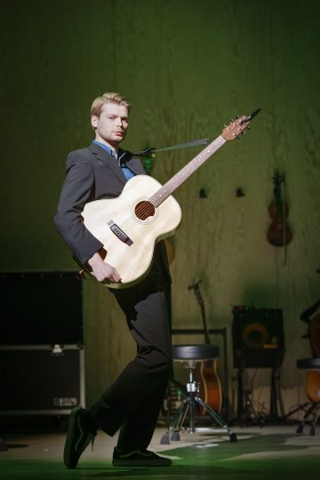 Lavalla seisoo pukuun ja lenkkareihin pukeutunut mies, joka soittaa akustista kitaraa. Taustalla on musiikkivälineitä, roikkuvia viuluja ja jakkara, ja vihreä valaistus luo dramaattista tunnelmaa.
