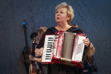 Lyhyet vaaleat hiukset omaava nainen soittaa lavalla punaista harmonikkaa, ja taustalla on muita muusikoita ja soittimia.