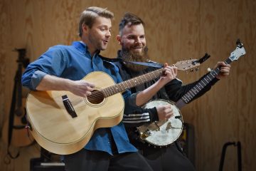 Kaksi muusikkoa, joista toinen soittaa akustista kitaraa ja toinen banjoa, esiintyvät innokkaasti yhdessä lavalla puista taustaa vasten. Molemmat näyttävät nauttivan musiikista.