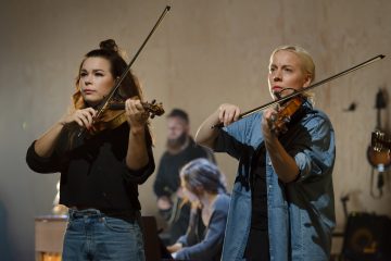 Etualalla kaksi naista soittaa viulua keskittyneenä esitykseensä. Taustalla näkyvät mies ja toinen nainen, joiden ympärillä on soittimia ja välineitä, mikä viittaa ryhmämusiikkisessioon tai harjoituksiin.