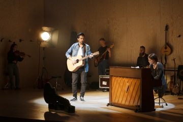 Mies soittaa akustista kitaraa ja laulaa lavalla naisen kanssa pianon ääressä, ja heitä säestävät viulisti, basisti ja heidän takanaan loistava valo hämärässä musiikkiesitysympäristössä.