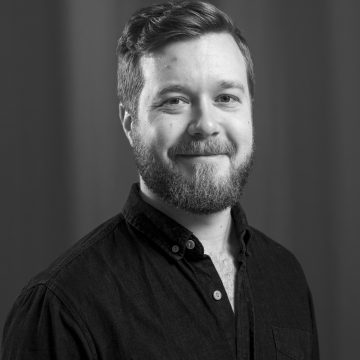 Parrakas, lyhythiuksinen mies, jolla on tumma nappipaita, hymyilee pehmeästi kameraan katsoen. Kuva on mustavalkoinen ja siinä on tavallinen, sumea tausta.