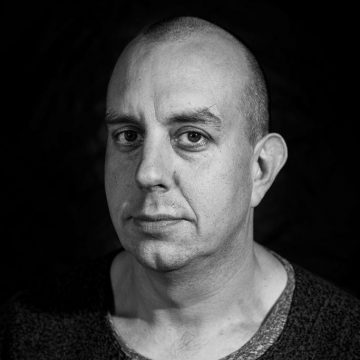 Mustavalkoinen muotokuva keski-ikäisestä miehestä, jolla on ajeltu pää, päällään kuvioitu pusero, ja joka katsoo suoraan kameraan neutraali ilme tummalla taustalla.