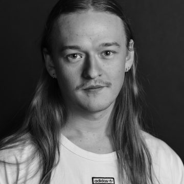 Mustavalkoinen muotokuva henkilöstä, jolla on pitkät hiukset, kevyet viikset ja korvakorut, yllään valkoinen Adidas t-paita, katselee suoraan kameraan neutraali ilme tummaa taustaa vasten.