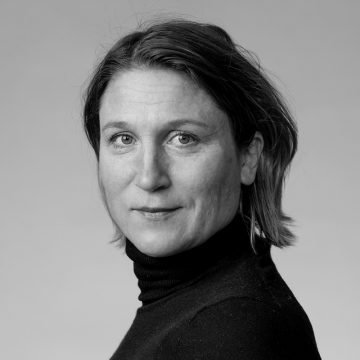 Mustavalkoinen muotokuva naisesta, jolla on vaaleat silmät ja olkapään mittaiset hiukset, päällään musta poolokaulus, katselee suoraan kameraan tavallista taustaa vasten.