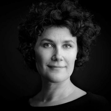 Mustavalkoinen muotokuva naisesta, jolla on lyhyet, kiharat hiukset ja tumma toppi. Hän katsoo suoraan kameraan lempeällä, neutraalilla ilmeellä tummaa taustaa vasten.