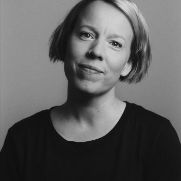 Mustavalkoinen muotokuva henkilöstä, jolla on lyhyet vaaleat hiukset, nenärengas ja tumma t-paita, katselee kameraan hymyillen, taustan ollessa tavallinen.