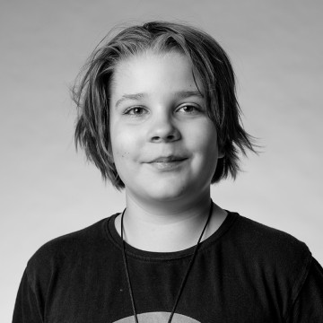 Mustavalkoinen muotokuva nuoresta henkilöstä, jolla on lyhyet, sekaiset hiukset, yllään tumma t-paita ja kaulakoru, ja joka hymyilee pehmeästi katsoen kameraan tavallista taustaa vasten.