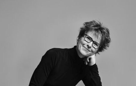 Lyhythiuksinen, silmälasipäinen henkilö, jolla on musta poolokaulus, hymyilee ja nojaa päätään käteensä vaaleaa taustaa vasten.