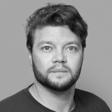 Mies, jolla on aaltoileva tukka ja parta, katsoo suoraan kameraan neutraali ilme kasvoillaan. Hänellä on yllään tumma paita, ja tausta on tavallinen ja vaalea.