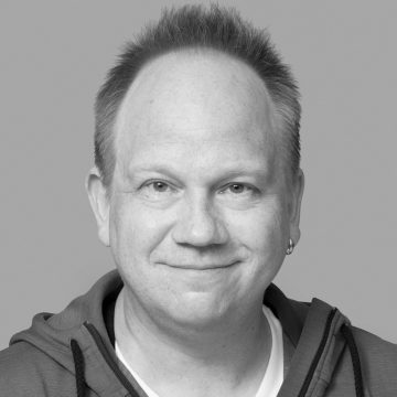 Keski-ikäinen mies, jolla on lyhyet vaaleat hiukset ja korvakoru, hymyilee kameralle. Hänellä on päällään huppari vaalean paidan päällä, ja tausta on yksivärinen ja harmaa.
