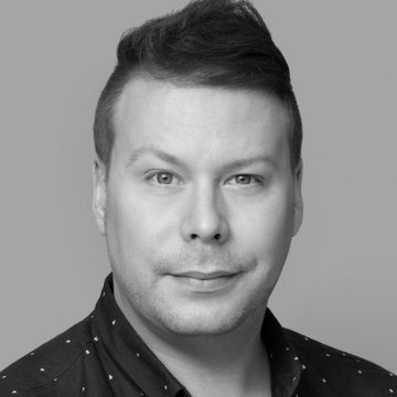 Mustavalkoinen muotokuva henkilöstä, jolla on lyhyet, tummat, sivulle tyylitellyt hiukset, päällään tumma, kuviollinen paita ja joka katsoo suoraan kameraan neutraali ilme.