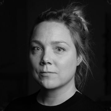 Mustavalkoinen muotokuva naisesta, jolla on vaaleat taakse vedetyt hiukset, katselee suoraan kameraan neutraali ilme kasvoillaan, yllään tumma toppi tummaa taustaa vasten.