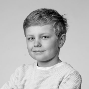 Nuori poika, jolla on lyhyet, vaaleat hiukset ja hento hymy, päällään vaalea pusero, poseeraa mustavalkoisessa muotokuvassa tavallista, vaaleaa taustaa vasten.