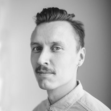 Mustavalkoinen muotokuva nuoresta miehestä, jolla on vaalea iho, lyhyet ylöspäin muotoillut hiukset, viikset ja korvakoru. Hänellä on kauluspaita ja hän katsoo hieman sivulle neutraali ilme kasvoillaan.