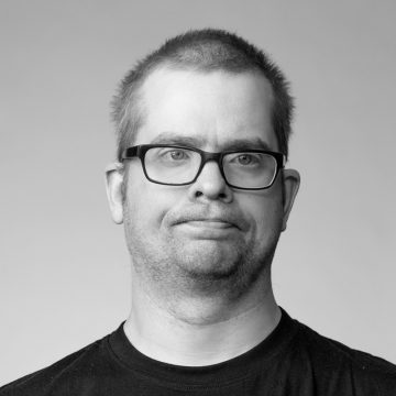 Lyhythiuksinen, silmälasipäinen mies, jolla on tumma t-paita, katsoo hieman kameran ulkopuolelle neutraalilla ilmeellä vaaleaa taustaa vasten.