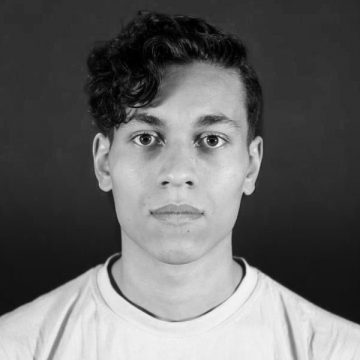 Nuori henkilö, jolla on lyhyet, kiharat tummat hiukset ja vakava ilme, on kasvot menosuuntaan tummaa taustaa vasten, ja hänellä on yllään vaalea paita.